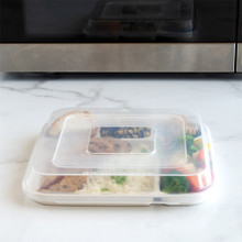 Nordic Ware aluminum divided dinner tray with clear lid, containing leftover food portions, positioned in front of microwave
