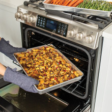 Hands removing Nordic Ware aluminum Big Batch Pan from oven containing roasted vegetables and meat