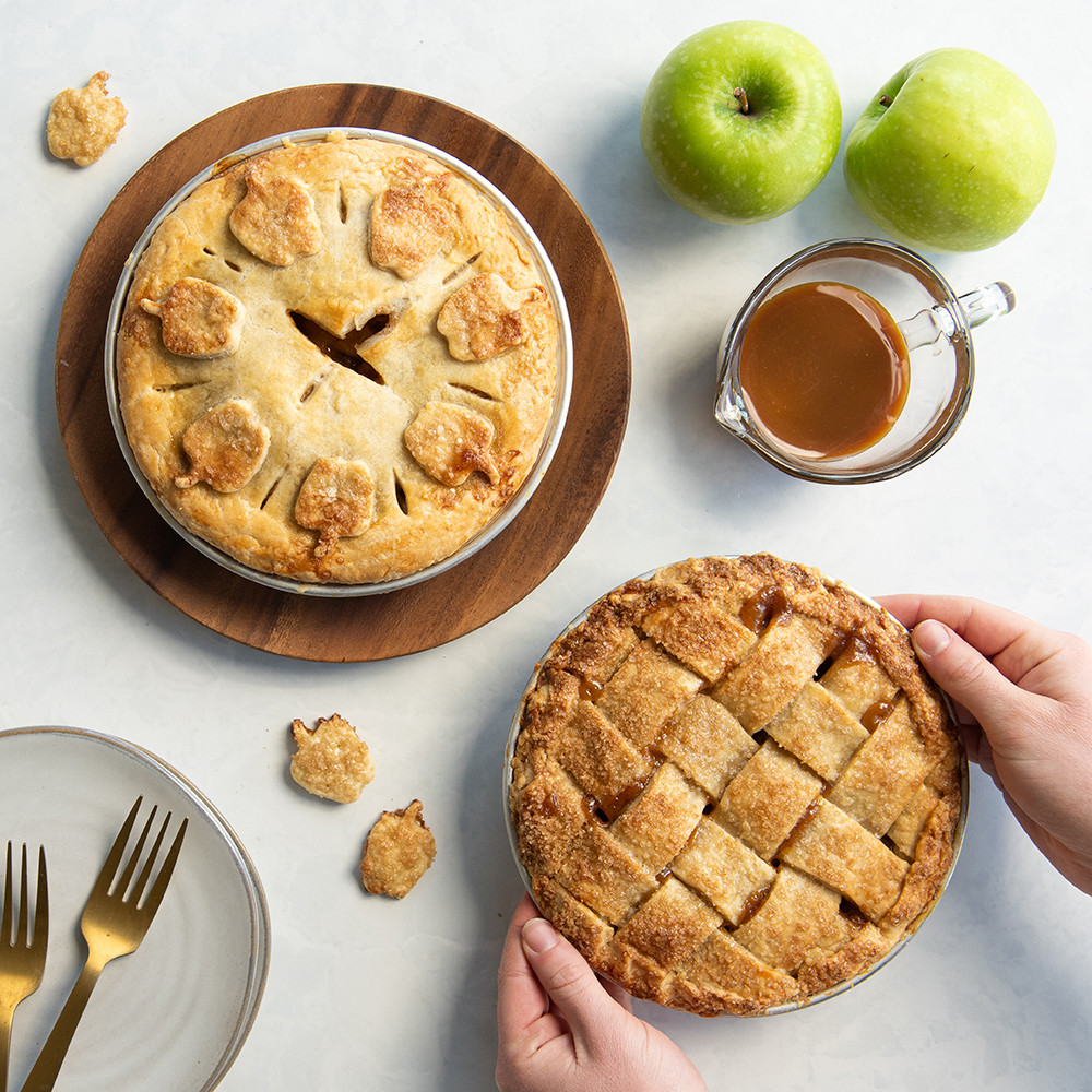 Two golden mini apple pies in Nordic Ware 5-inch aluminum pie pans, one with lattice crust, one with leaf cutouts