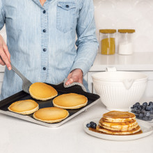 White Nordic Ware Better Batter Bowl with pour spout next to golden pancakes cooking on griddle