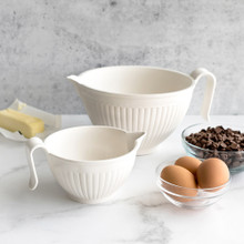 Nordic Ware white Better Batter Bowl with spout alongside mixing bowls filled with flour, eggs, and baking ingredients