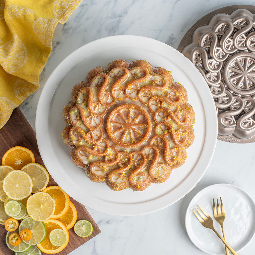 glazed citrus twist cake on white cake stand with sliced lemons, and small plate with forks