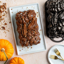 Overhead image of glazed Sunflower Pumpkin Loaf cake sprinkled with pecan pieces on white plate surrounded pan, pumpkins and crushed pecans