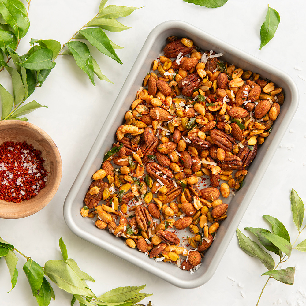Nordic Ware aluminum eighth sheet pan with mixed nuts, small bowl of spices, and green leaves arranged overhead