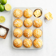 Nordic Ware aluminum muffin pan with golden-brown lemon poppy seed muffins, fresh limes and coconut flakes