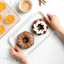 Two golden mini bundt cakes on white plate with orange slices, cinnamon bowl, and cooling rack in background