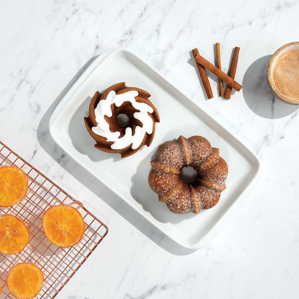 Two golden bundt cakes on plates with orange slices on cooling rack and bowl of cinnamon spice