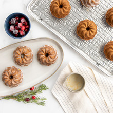 Golden baked pirouette bundtlettes with swirled ridged design on white plate next to Nordic Ware aluminum pan