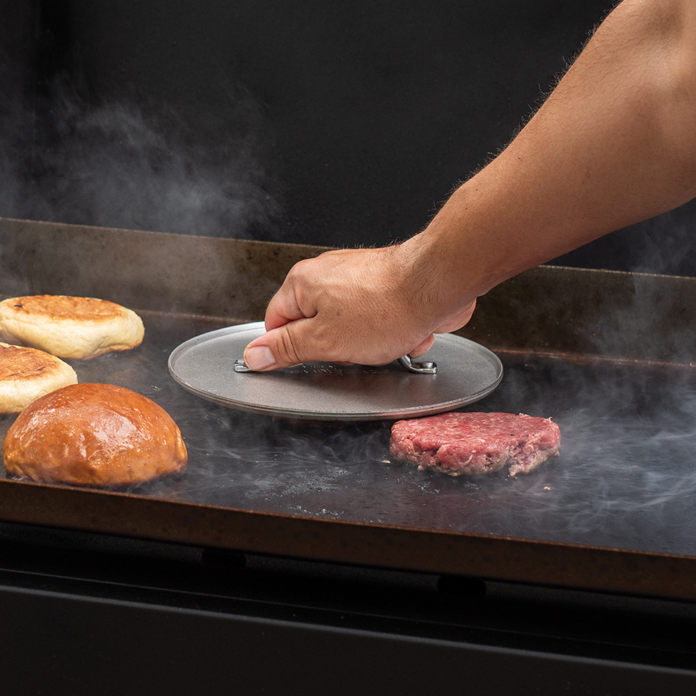 Nordic Ware aluminum grill press compressing raw beef burger patty on grill grates, silver heavyweight design