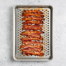 Nordic Ware aluminum high-sided half sheet pan with wire cooling rack, holding strips of cooked bacon