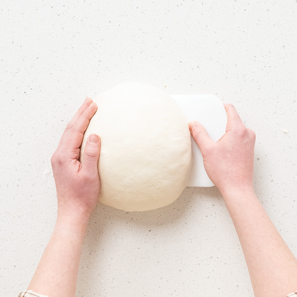 Stainless steel bench scraper lifting shaped sourdough dough from floured wooden countertop surface