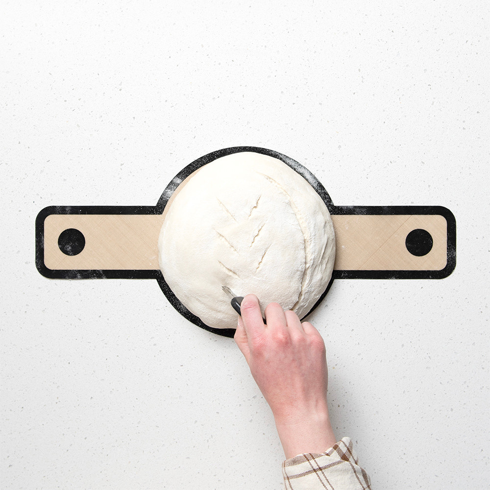 Raw sourdough dough on parchment sling being scored with Nordic Ware lame cutter, creating diagonal slashes