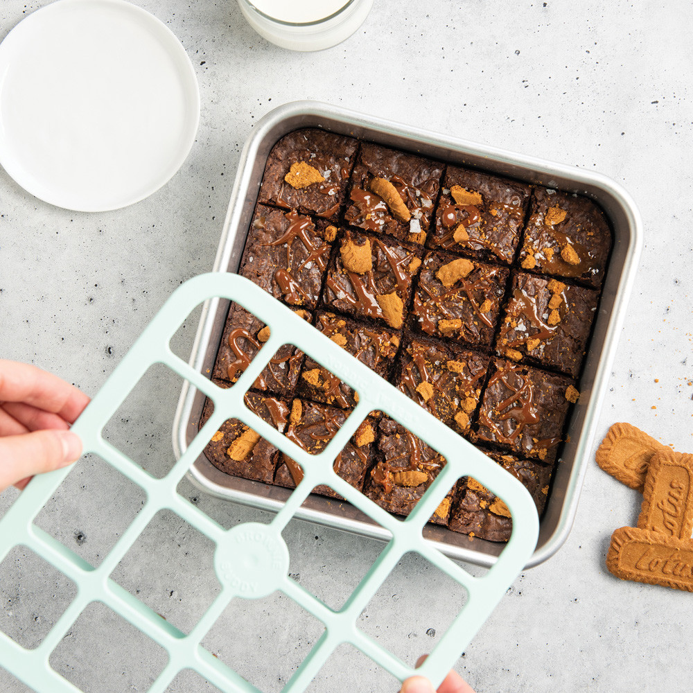 Nordic Ware Brownie Buddy metal cutter tool held over square pan of cut chocolate brownies with milk glass