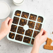 Hands using Nordic Ware brownie cutter tool over square pan of baked brownies with milk and cookies nearby