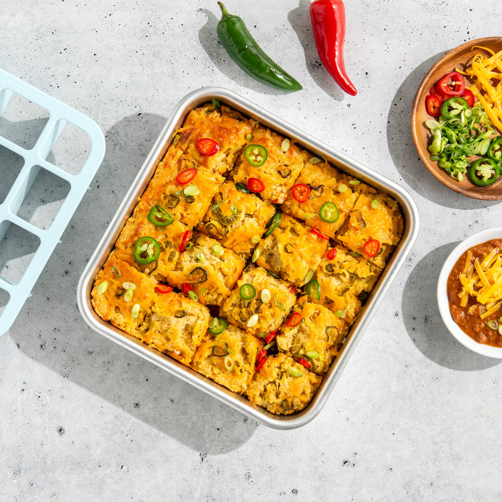 Nordic Ware square aluminum baking pan with cut taco casserole squares, red brownie cutter tool beside pan
