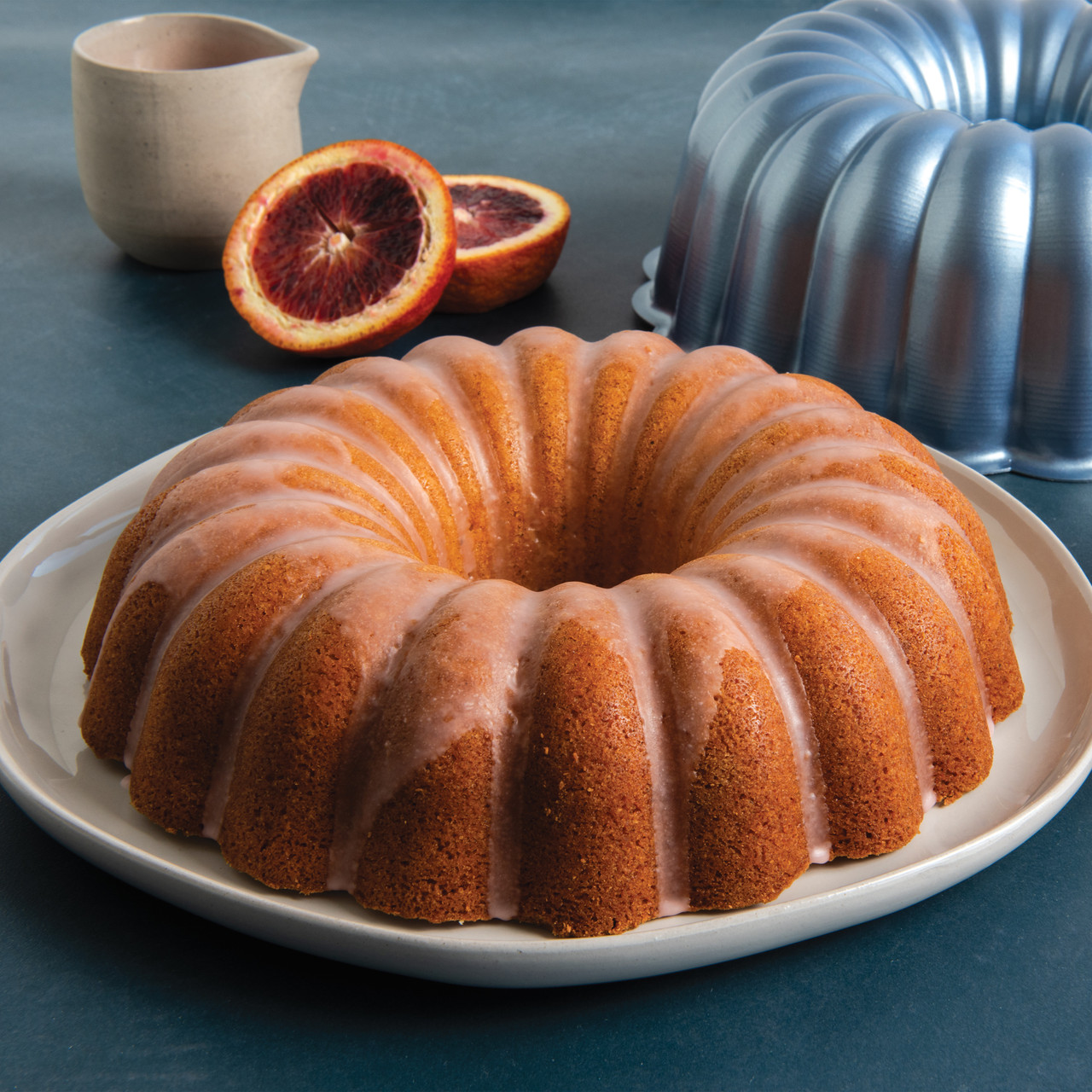 Nordic Ware Formed Elegant Party Bundt pan with golden baked cake, fluted design, mango slices and icing nearby