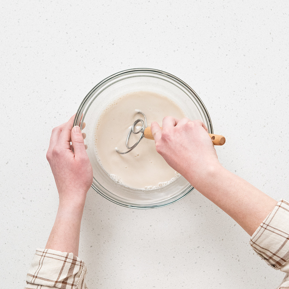 Hands using Nordic Ware wooden Danish whisk to mix sourdough dough in clear glass mixing bowl