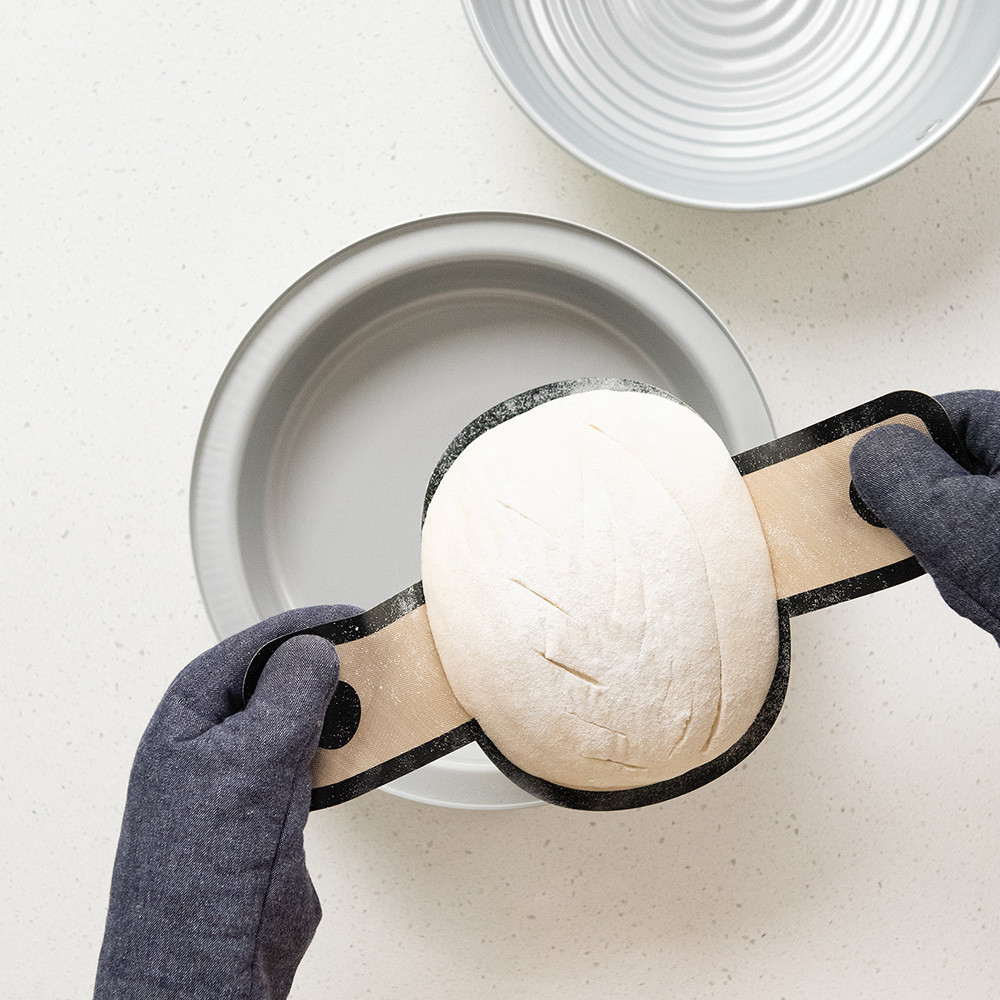 Hands placing shaped sourdough dough into Nordic Ware aluminum loaf pan using parchment paper sling