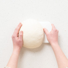 Stainless steel bench scraper lifting shaped sourdough dough from floured countertop surface