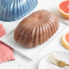 Glazed fluted loaf cake beside Nordic Ware aluminum fluted loaf pan with orange slices on white countertop