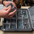 Person organizing screws and hardware inside a UbeCube (modular-storage-crate) BaseBox (hardware-organizer-box) using removable divider bins in a workshop setting.