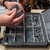 Person organizing screws and hardware inside a UbeCube (modular-storage-crate) BaseBox (hardware-organizer-box) using removable divider bins in a workshop setting.