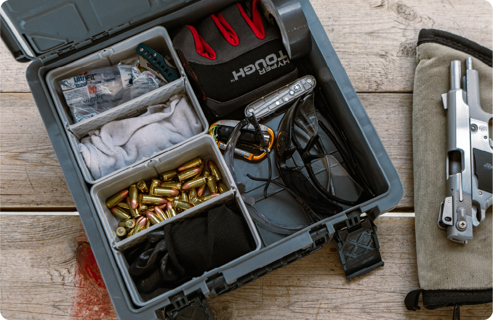 Open CrateShot Basebox™ organized with ammunition, gloves, safety glasses, medical supplies, and range gear on a wooden surface.