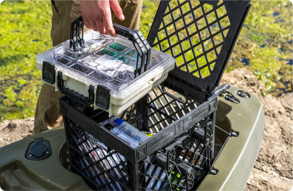 Person lifting a clear-lid BaseBox from a black UbeCube crate on a kayak by the water.