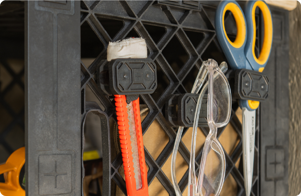Close-up of the UbeCube Plier Holder quarter-turn accessory mounts holding a utility knife, safety glasses, and scissors on a black crate side grid.