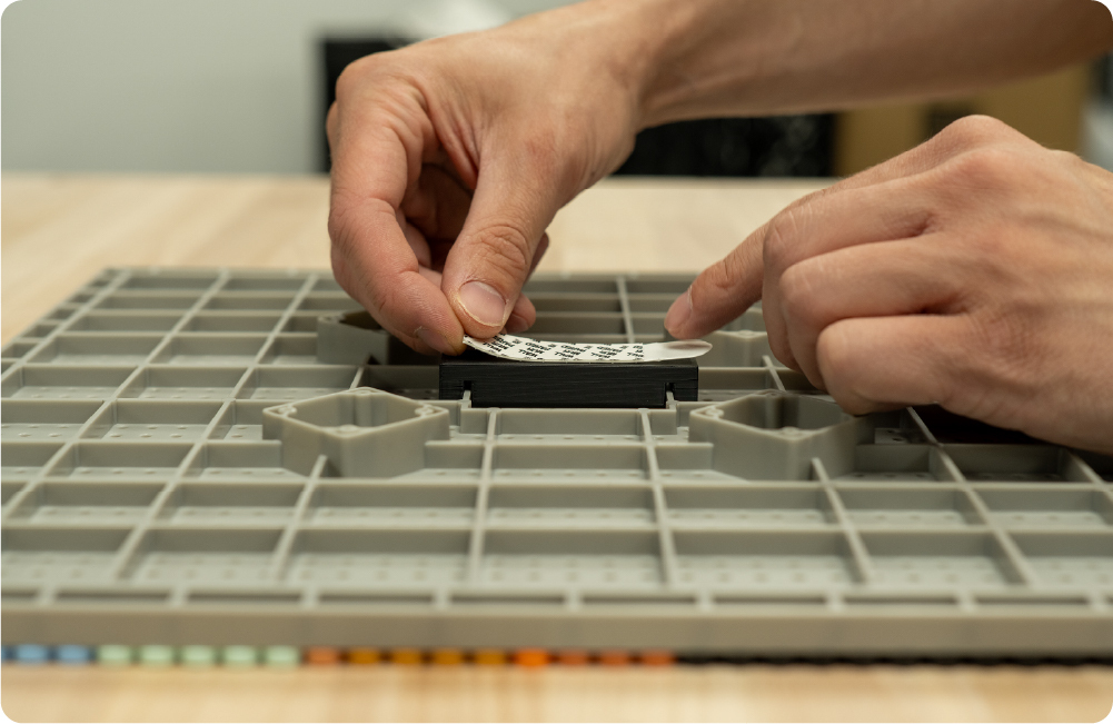 Hands applying a 3M Sticky pad to the back of a gray Pixel Panel build plate.
