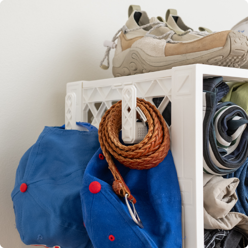 White UbeCube CrateCloset with white CrateHooks holding hats and a braided belt beside rolled clothing and shoes.