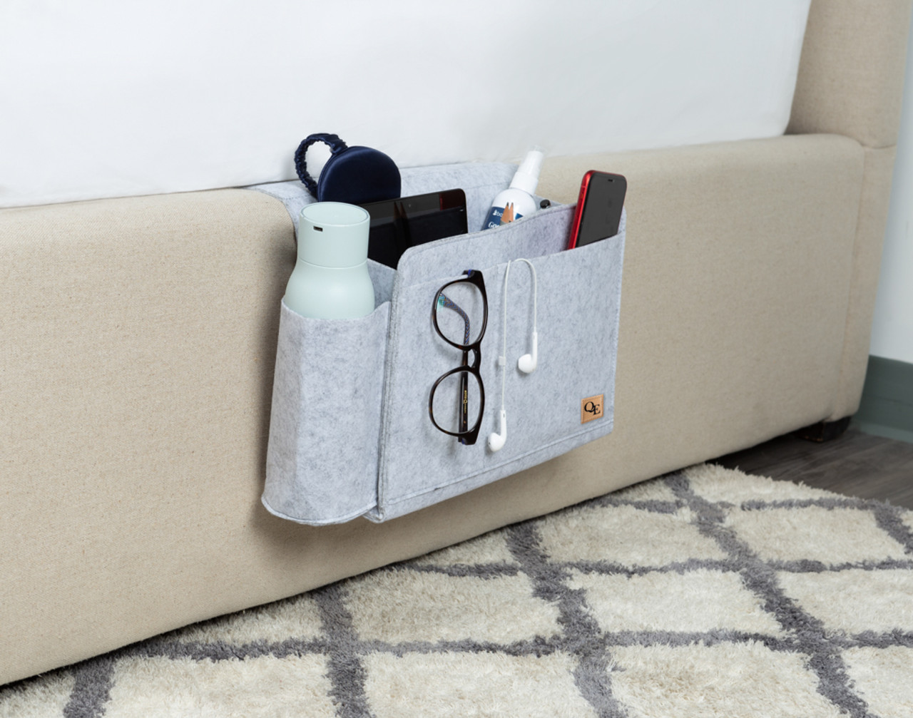Angled view of our Bedside Felt Pocket in Light Grey hanging off the edge of a bed with glasses, a water bottle, and more inside.