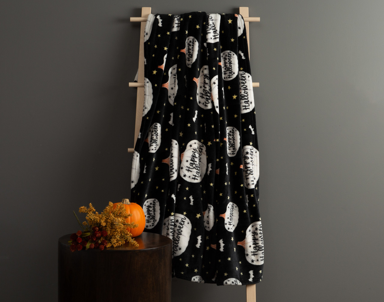 Our Happy Halloween Fleece Throw hanging from a ladder in a dark room, beside a small table with a pumpkin.