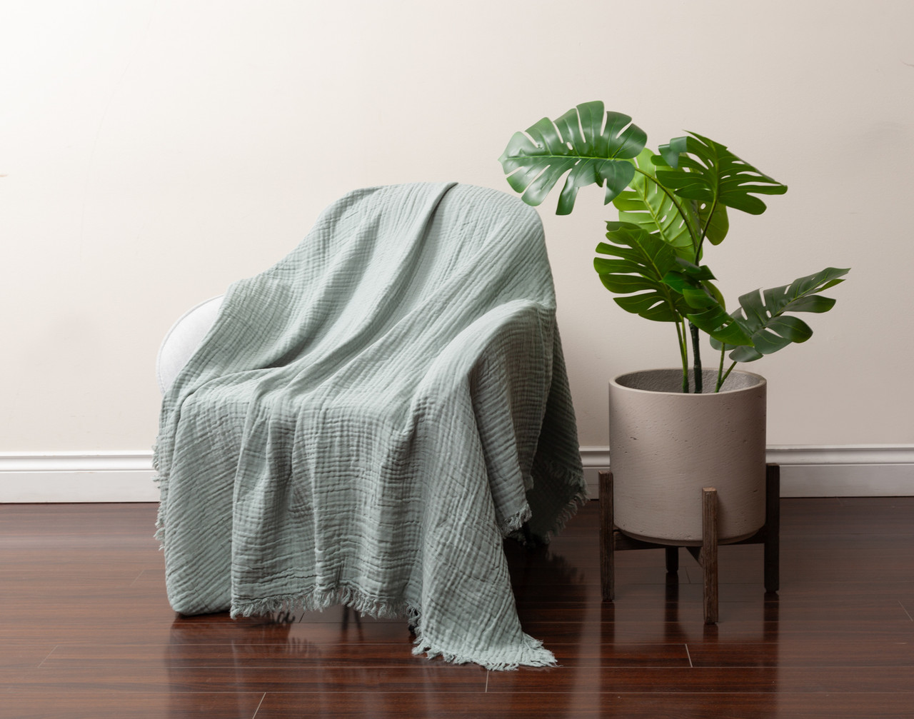 Angled view of our Muslin Gauze Throw in Seagrass draped over a chair next to a tall leafy plant.