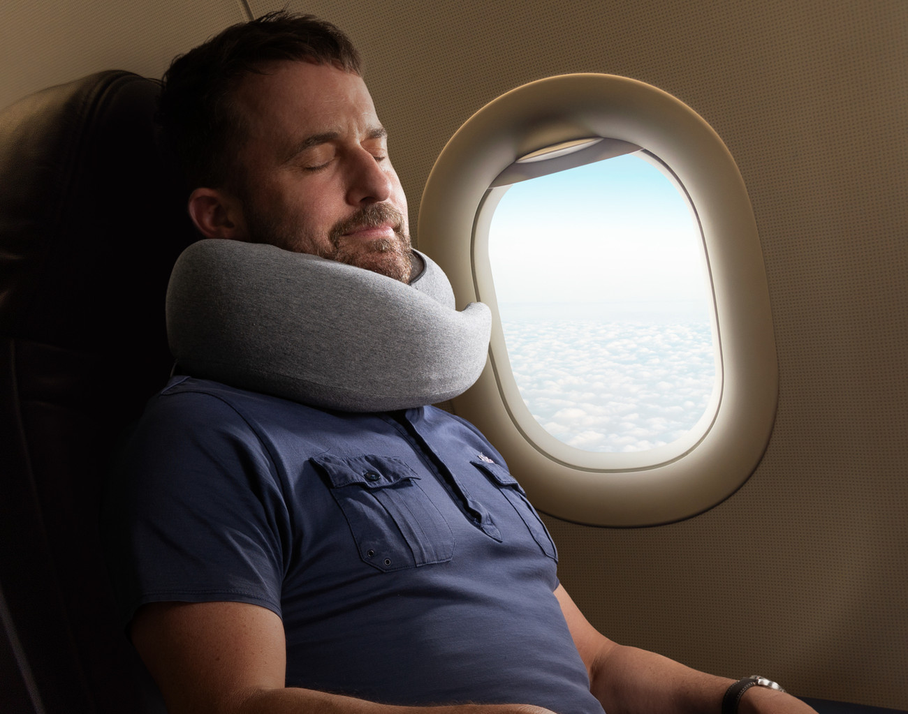 Homme barbu dormant dans un avion et portant notre oreiller de voyage Neck-Hug à côté d'une fenêtre ouverte.