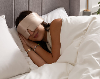A woman in bed wearing our 100% Mulberry Silk Blackout Sleep Mask in Champagne.