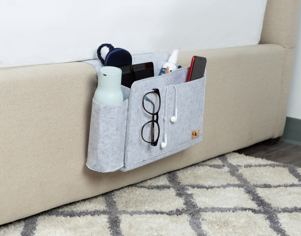 Angled view of our Bedside Felt Pocket in Light Grey hanging off the edge of a bed with glasses, a water bottle, and more inside.