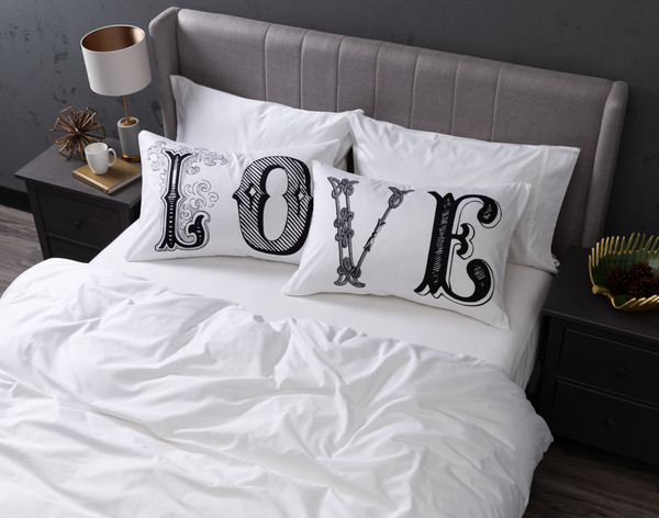 Vue aérienne de nos taies d'oreiller Love Pillow Talk habillées sur des oreillers Queen dans un grand lit blanc.