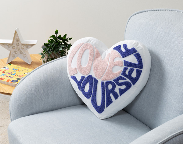Vue en angle de notre coussin Love Yourself BeaYOUtiful en forme de cœur, posé sur un petit fauteuil gris.