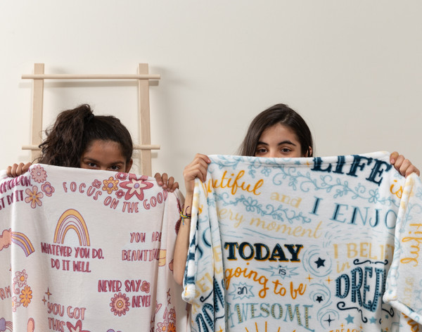 Two young girls holding up our BeaYOUtiful Throws side-by-side, including our You are Beautiful design (left) and our Life is Beautiful design (right).