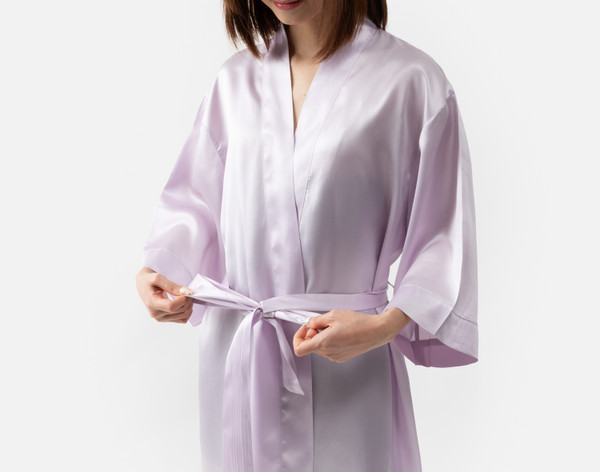 Gros plan de notre robe kimono en soie lavande portée par une jeune femme qui ajuste la ceinture coordonnée.