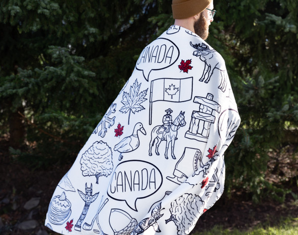 Back view of a person wearing our Welcome To Canada Fleece Throw as a cape outside among deep green trees.