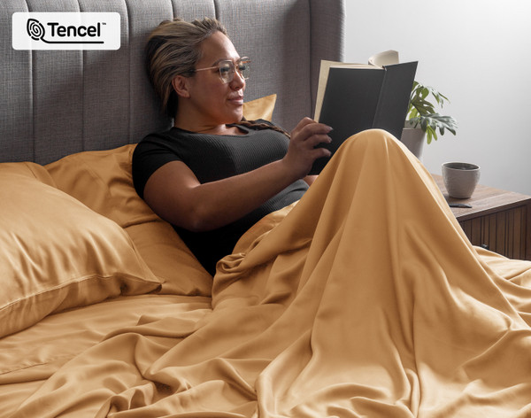 Woman sitting upright in a bed reading her book curled up in the flat sheet of our BeechBliss TENCEL™ Modal Sheet Set in Gilded.