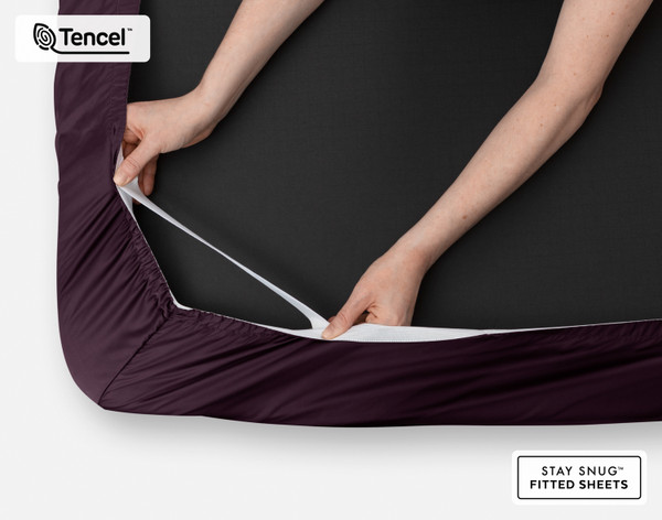 Close-up on two hands stretching the elastic snug fit band on the corner of our Eucalyptus Luxe TENCEL™ Lyocell Fitted Sheet in Purple.