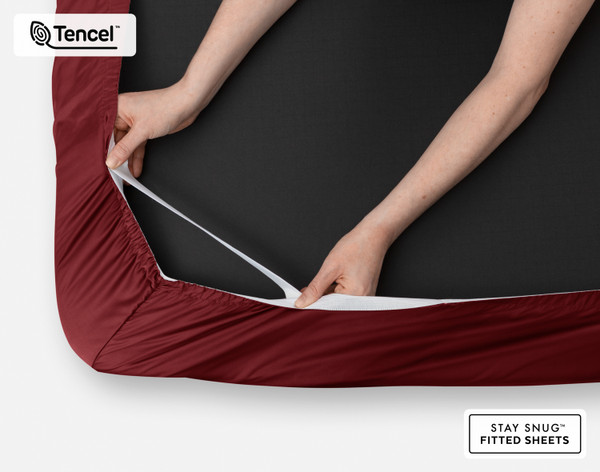Close-up on two hands stretching the elastic snug fit band on the corner of our Eucalyptus Luxe TENCEL™ Lyocell Fitted Sheet in Black Cherry.
