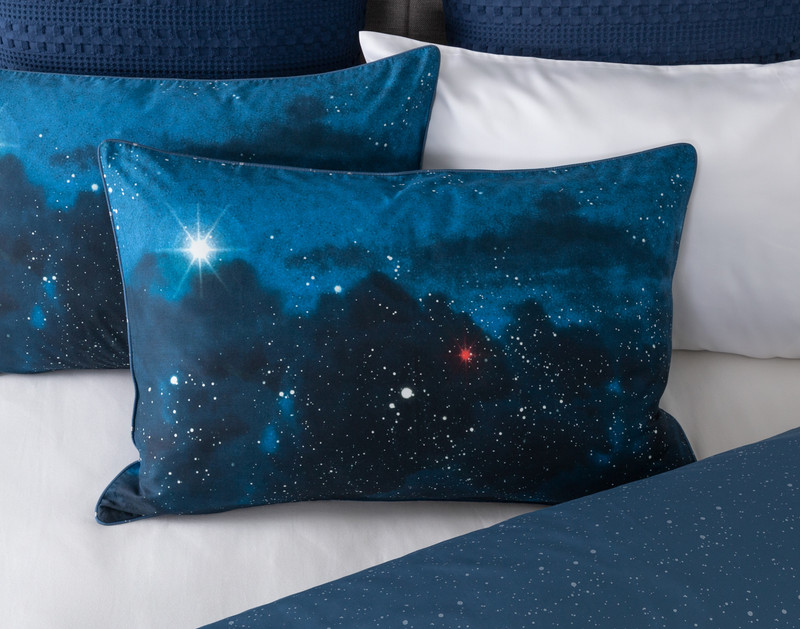 Front view of our Supernova Pillow Sham sitting against a matching pillow sham and white sheets to show its starry sky pattern.