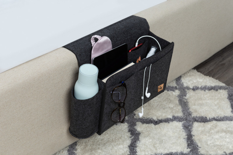 Vue latérale de notre pochette en feutre de chevet en gris foncé avec des objets essentiels du salon comme une télécommande, une tablette et une bouteille d'eau.