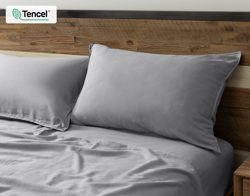 Vue en angle de nos Taies d'oreiller en chanvre gris posées contre une tête de lit en bois dans un lit habillé avec draps.