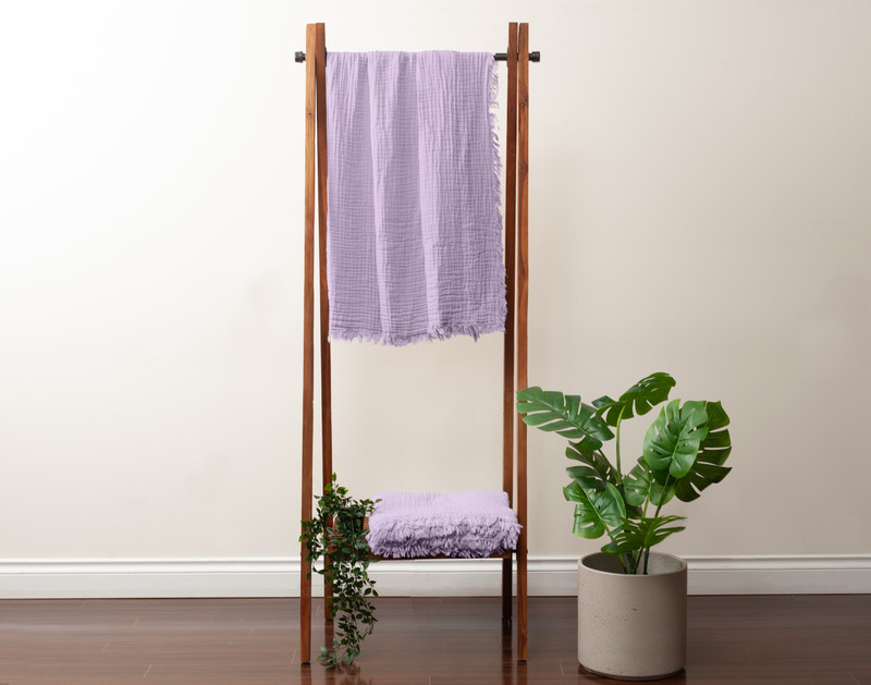 Front view of our Muslin Gauze Throw in Violet hanging on a tall fixture next to a short leafy plant.