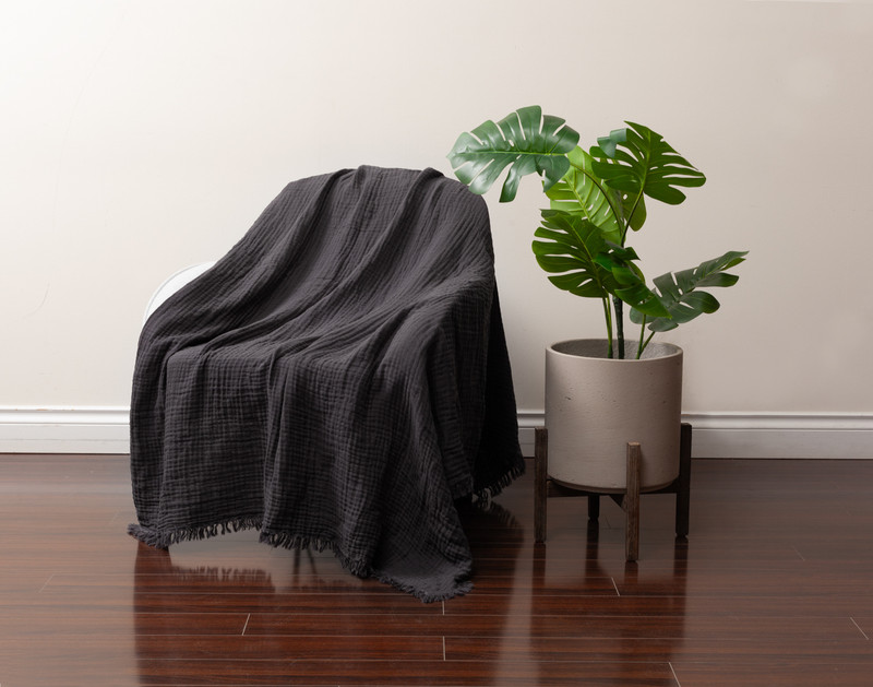 Angled view of our Muslin Gauze Throw in Obsidian draped over a chair next to a tall leafy plant.